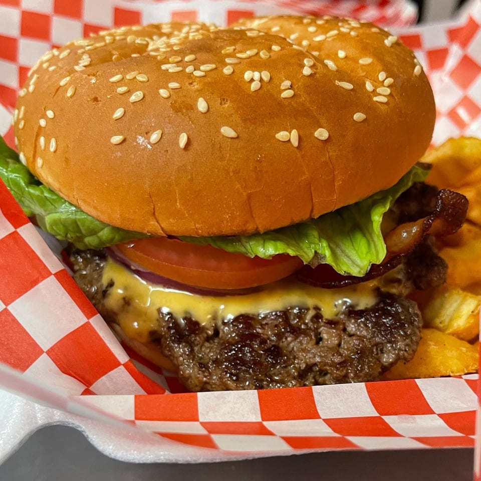 Cheeseburger with lettuce, tomato, and bacon, served with fries in a red and white checkered basket.