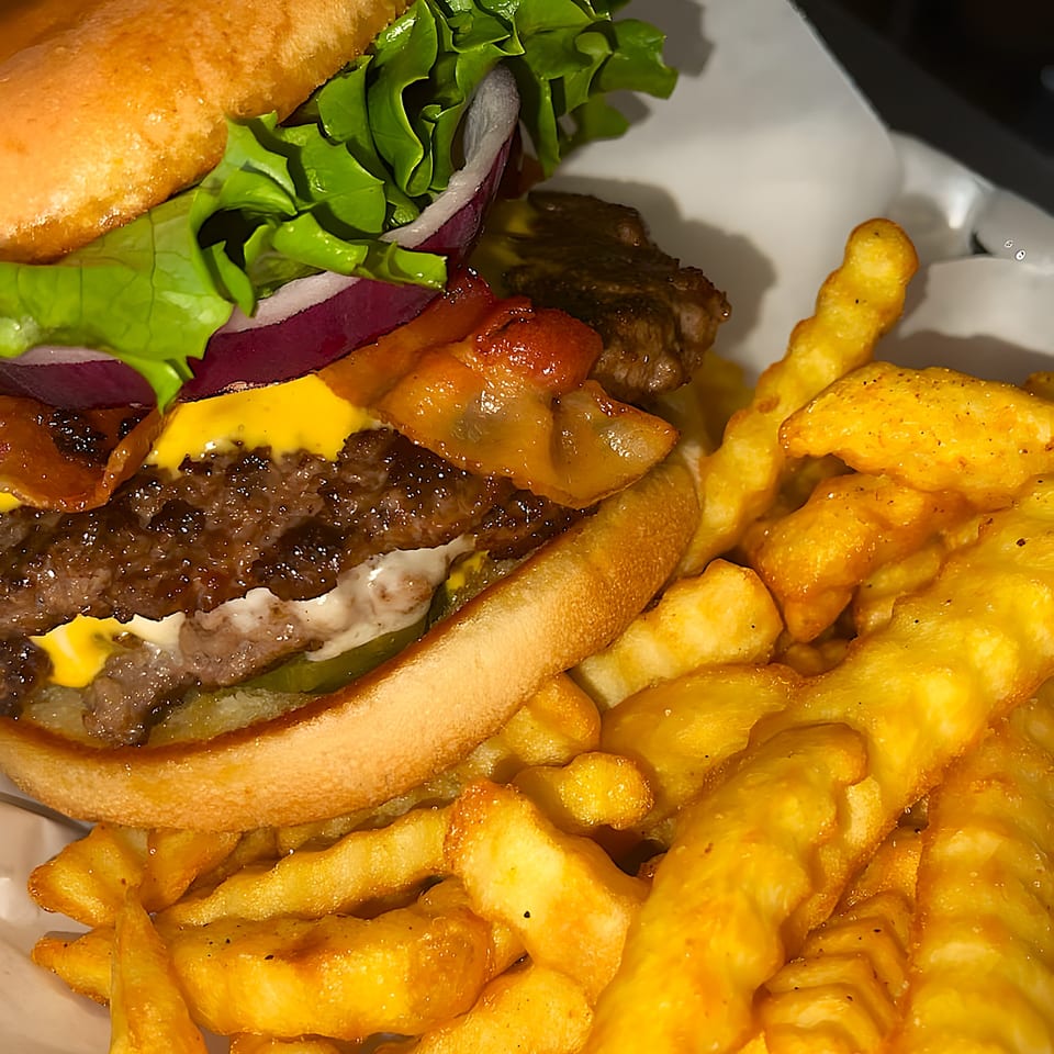 Close-up of a double cheeseburger with bacon, lettuce, onion, and pickles, served with crinkle-cut fries.