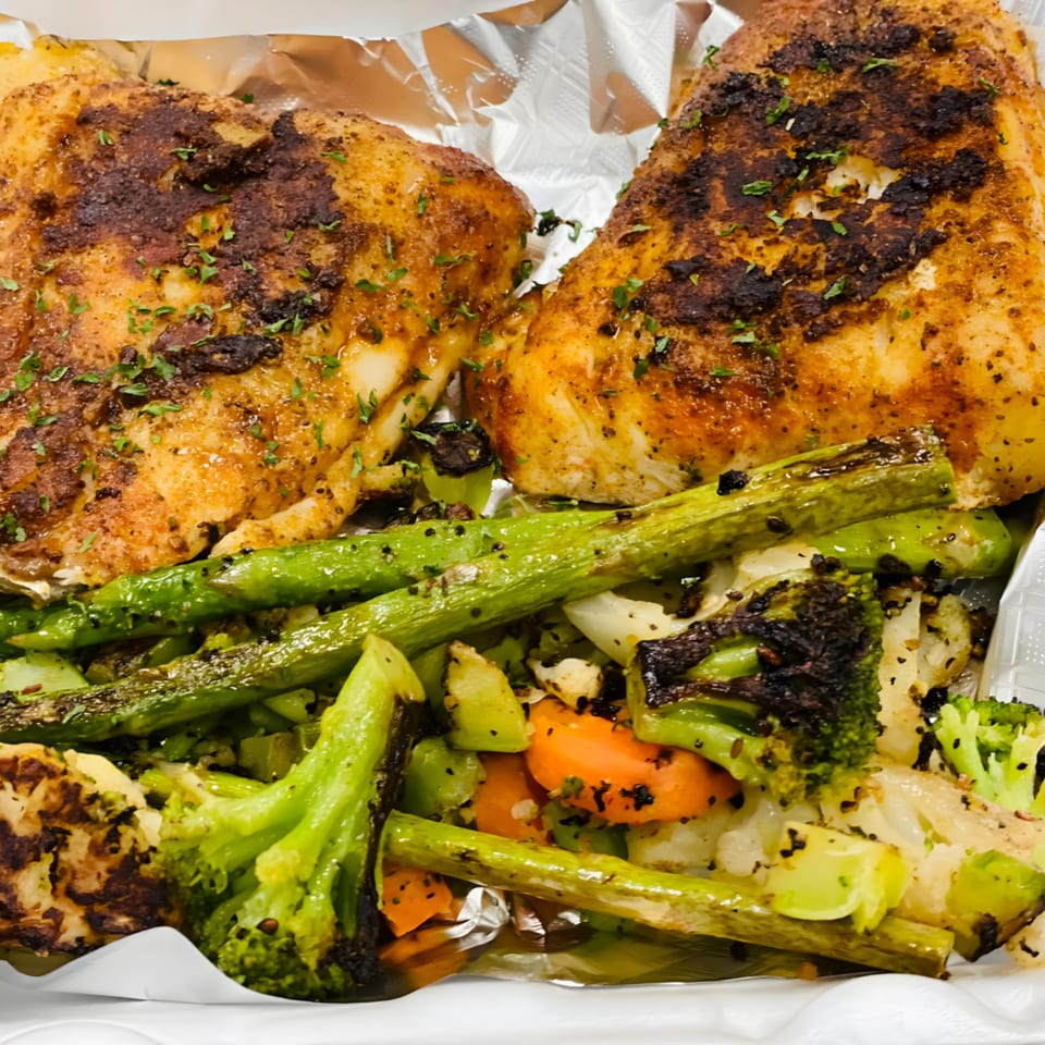 Two seasoned chicken breasts with roasted asparagus, broccoli, carrots, and cauliflower in a takeout container.