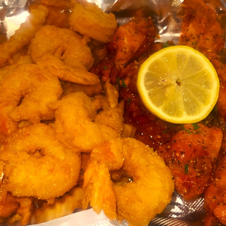 Golden fried shrimp and spicy chicken wings with a lemon slice, served in a foil container.
