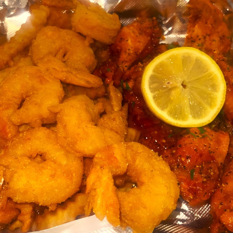 Golden fried shrimp and spicy chicken wings with a lemon slice, served in a foil container.