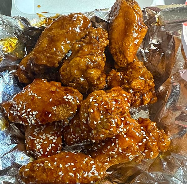 Close-up of sticky, glazed chicken wings with sesame seeds in a foil-lined takeout container.