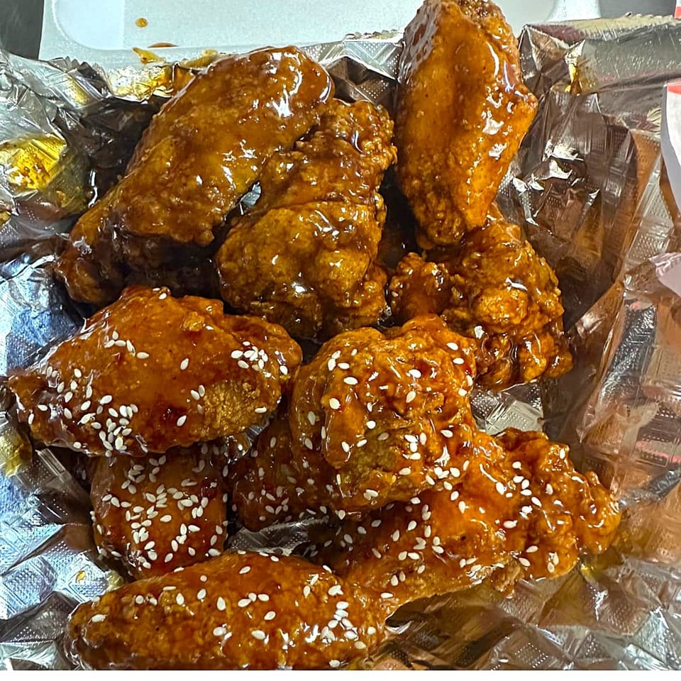 Close-up of sticky, glazed chicken wings with sesame seeds in a foil-lined takeout container.