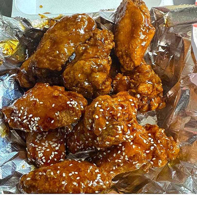 Close-up of sticky, glazed chicken wings with sesame seeds in a foil-lined takeout container.